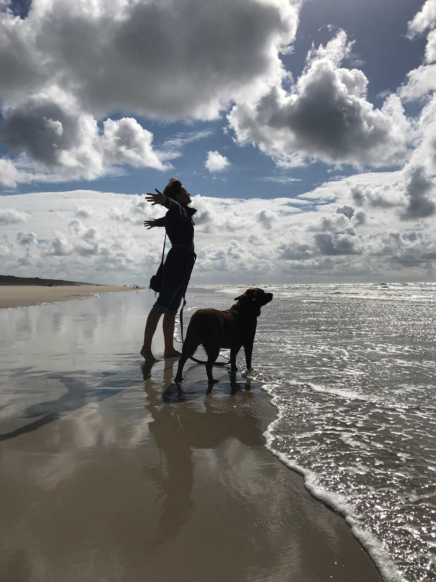 Katrin und ihre Hündin Fina stehen nebeneinander am Strand und blicken gemeinsam aufs Meer, die Arme weit ausgestreckt – Symbol für Verbundenheit, Vertrauen und ihren gemeinsamen Lebensweg