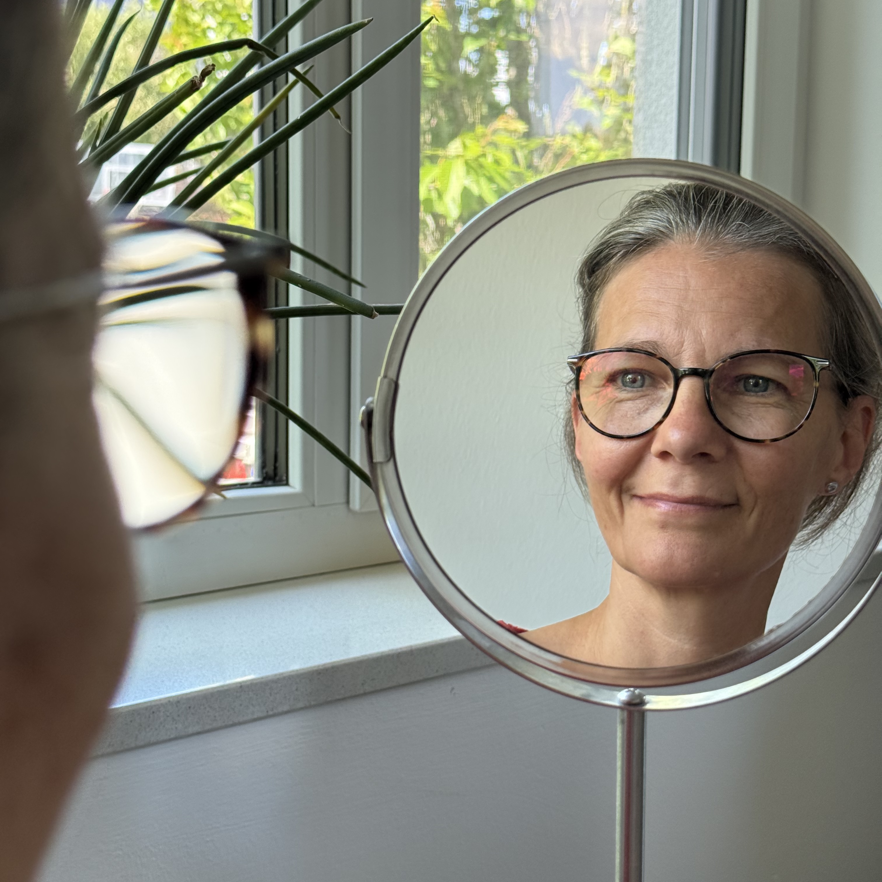 Katrin Strabel von Kafina Coaching blickt lächelnd in einen runden Standspiegel am Fenster – Symbol für den liebevollen und mutigen Blick auf sich selbst