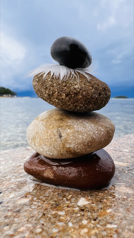 Übereinandergestapelte Steine mit einer Feder unter dem obersten Stein vor verschwommenem Meer, als Symbol für innere Balance, sanfte Impulse und Vertiefung auf dem persönlichen Weg.