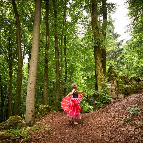 Frau mit weitschwingendem Rock tanzt fröhlich, lebendig und bewusst im Einklang mit sich selbst im Wald zwischen den Bäumen als Symbol für Wachstum, innere Freiheit und Muttersein gepaart mit Persönlichkeitsentwicklung .