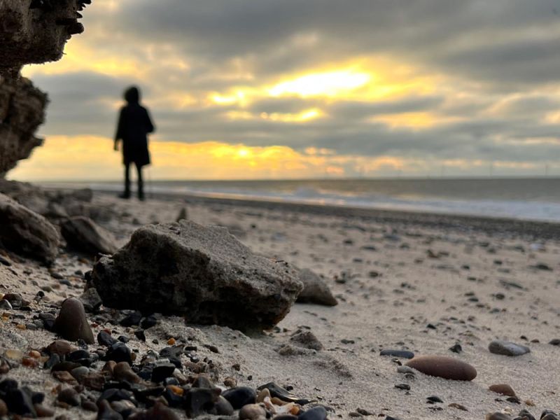 Symbolbild für emotionale Blockaden: Person am Meer mit bröckelnder Sandkante als Metapher für emotionalen unsichtbaren Ballast, der unser Gleichgewicht aus der Balance bringen kann.