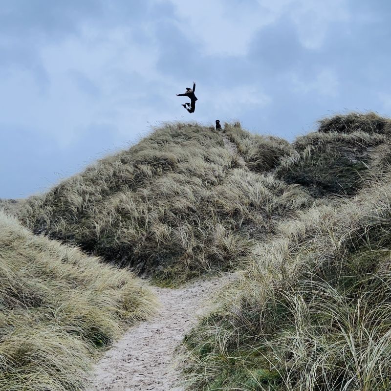 Eine Person springt freudig auf einer Düne in den bewölkten Himmel – Symbol für Leichtigkeit, Aufbruch und die Freude nach der inneren Transformation