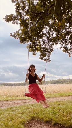 Katrin Strabel sitzt im Sommer mit einem langen, schwingenden Rock auf einer Baumschaukel an einem kräftigen Baum. Ihr freudiges Lachen erinnert an kindliche Leichtigkeit und unbeschwerte Lebensfreude.
