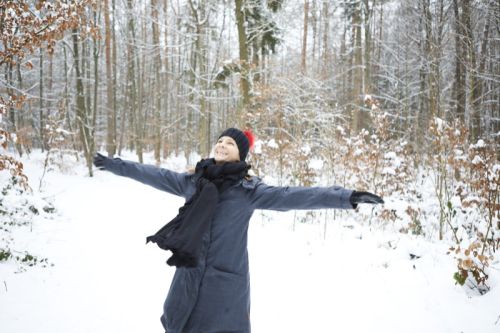 Katrin Strabel steht im tief verschneiten Winterwald, dreht sich mit ausgestreckten Armen und lacht frei und glücklich. Das Bild steht für Leichtigkeit, innere Freiheit und Lebensfreude.