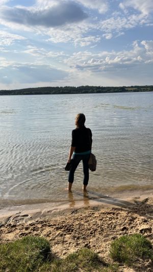 Katrin Strabel steht mit dem Rücken zur Kamera am Seeufer, die Knöchel im Wasser, und blickt über den See in die Ferne. Das Bild vermittelt Ruhe, Weite und einen bewussten Moment des Innehaltens.